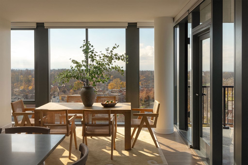 A table with chairs and a vase of flowers is in front of a window with a view of trees.