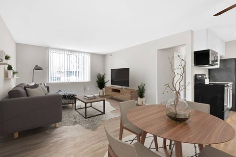 A modern living room with a grey sofa, a wooden coffee table, a TV, a refrigerator, and a wooden dining table with chairs.