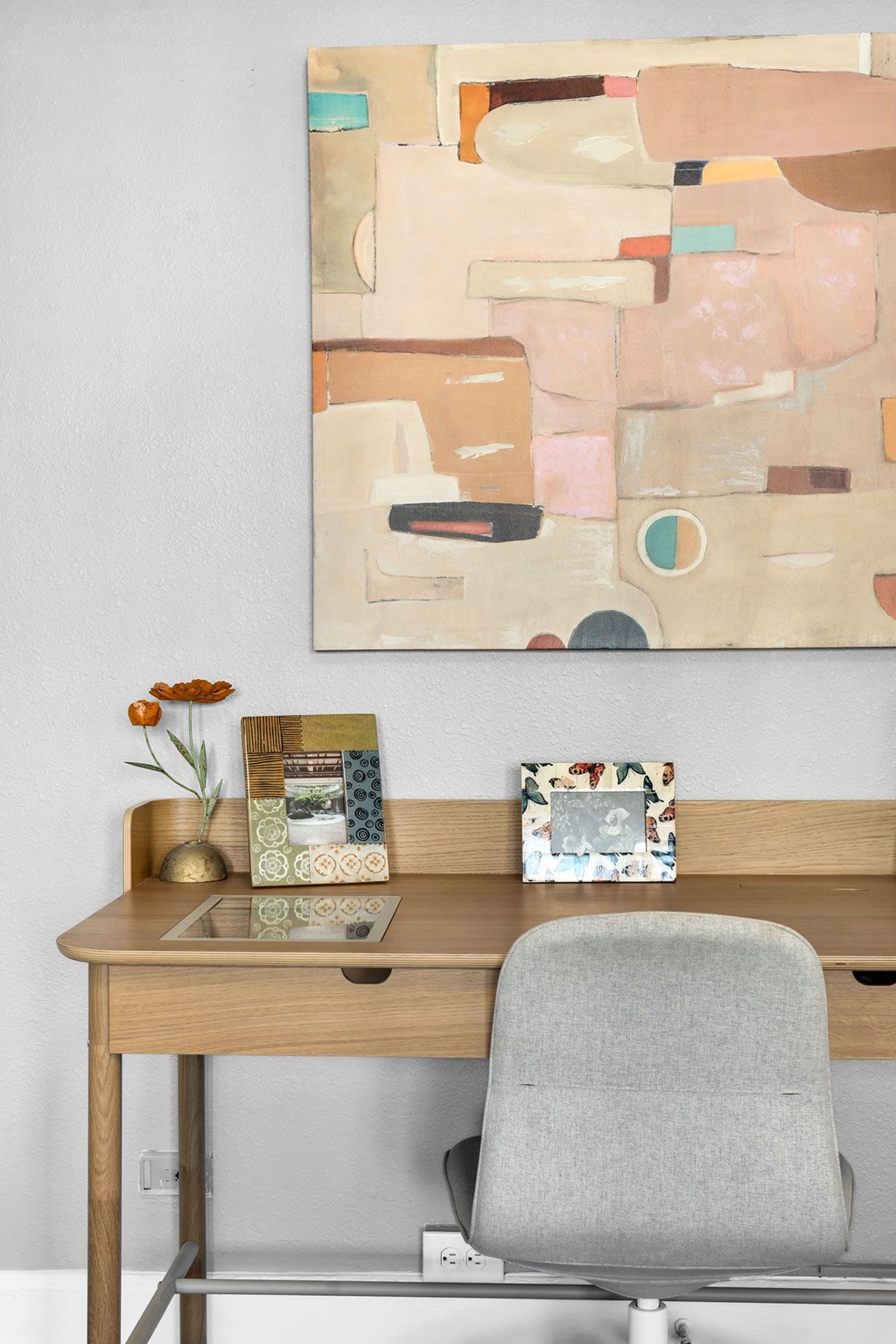 A painting hangs on a wall above a desk with a chair and some picture frames.