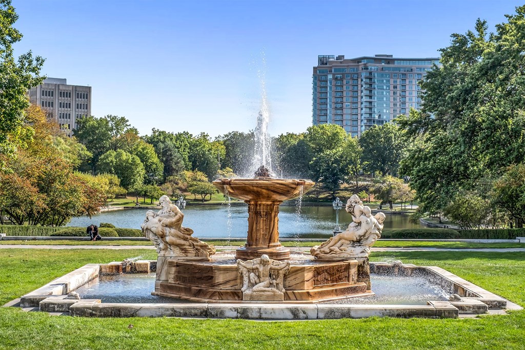 A fountain in the middle of a park with trees and buildings in the background.