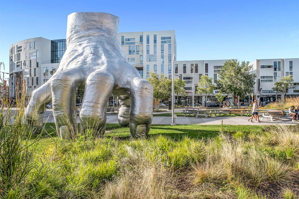 A large sculpture of a hand is in the foreground of a grassy area with buildings in the background.