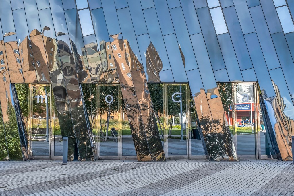 A large, modern sculpture made of mirrored panels is reflected in a glass panel.