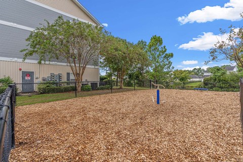 A playground with a blue post in the middle of a brown surface surrounded by trees and a building in the background.