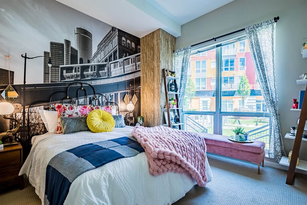 A bedroom with a bed, a window with a view of a city, and a pink blanket.