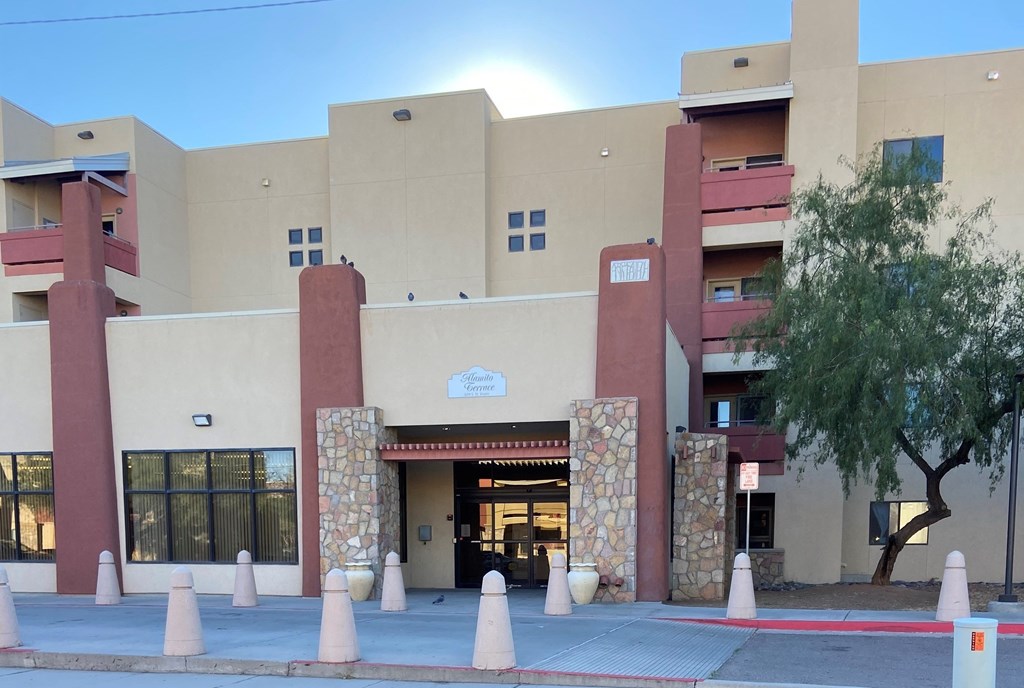 The image shows a building with a beige facade and a red entrance, surrounded by a row of trees.