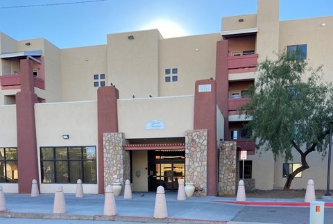 The image shows a building with a beige facade and a red entrance, surrounded by a row of trees.