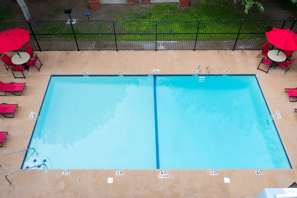 A rectangular pool with a blue tint is surrounded by a black fence and has red umbrellas on the side.