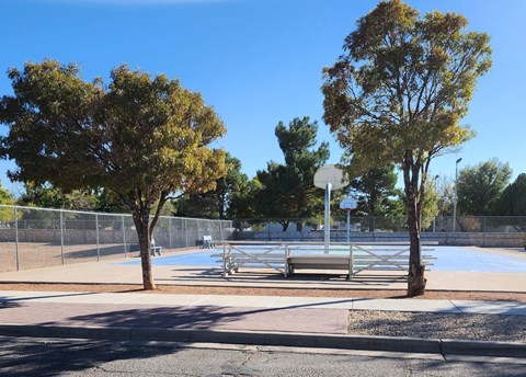 A park with a bench, a basketball court and a fence.
