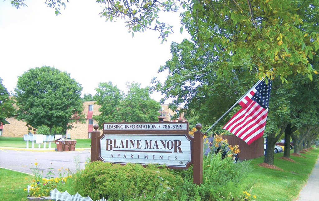 Blaine Manor Apartments sign with American flag, surrounded by trees, flowers, and a well-maintained lawn.