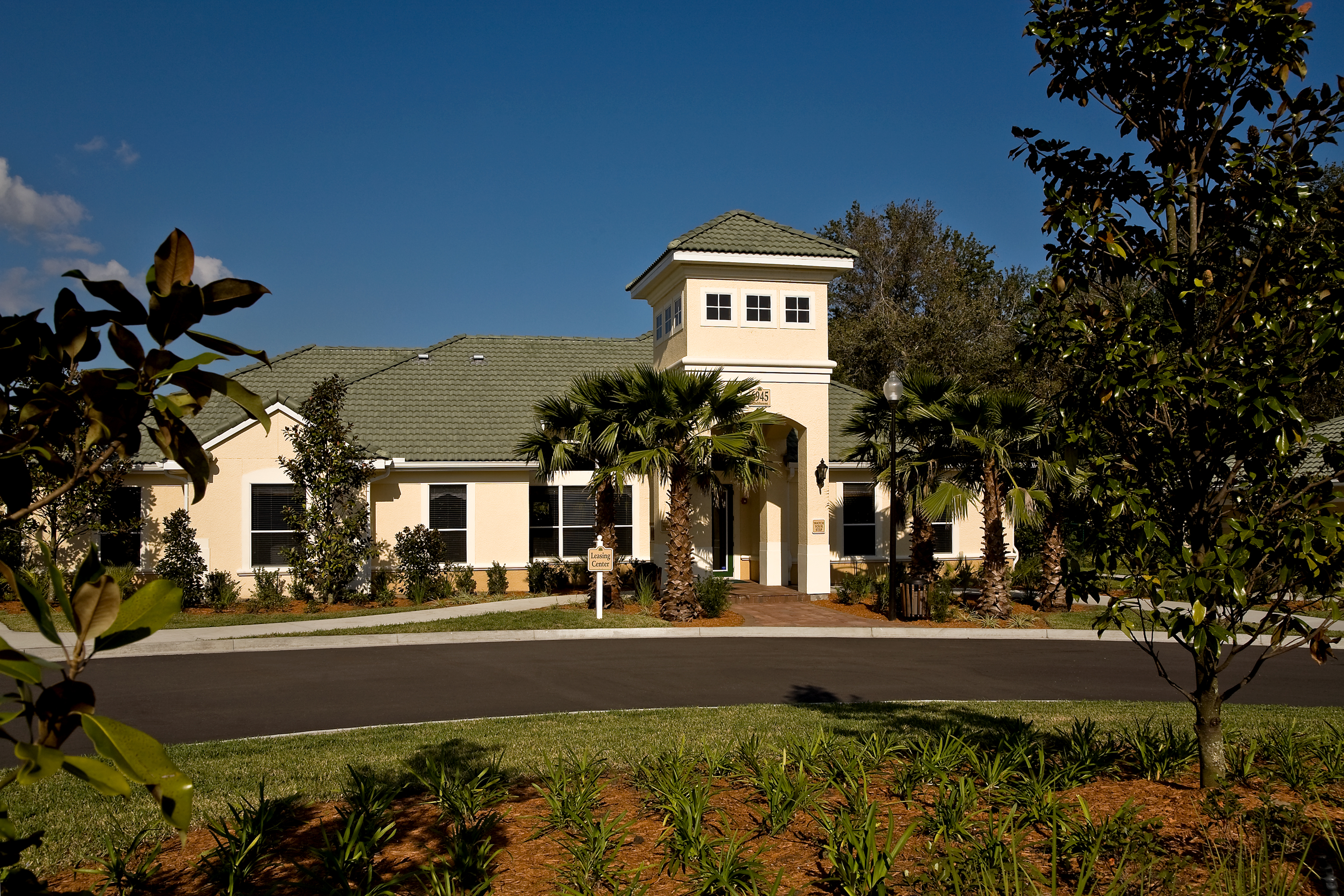 A white house with a green roof and a tower is surrounded by trees and shrubs.