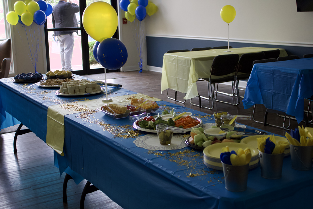 Community event setup with tables of food, balloons, and decorations in a blue and yellow theme