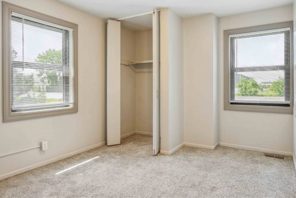 A room with a carpeted floor, two windows with blinds, and a closet with a shelf.