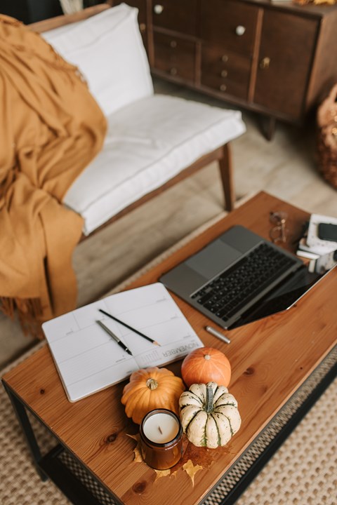 A wooden table with a laptop, a notebook, a pen, a pumpkin, and a candle on it.
