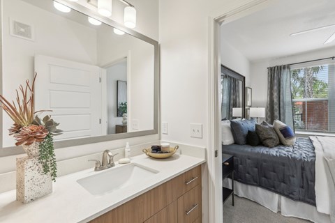 A modern bathroom with a large mirror, sink, and a bedroom in the background.