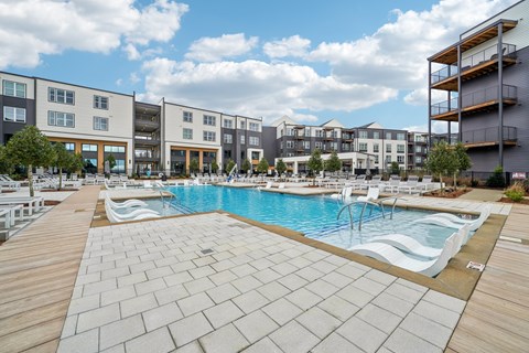 A large outdoor swimming pool with a wooden deck in front of apartment buildings.