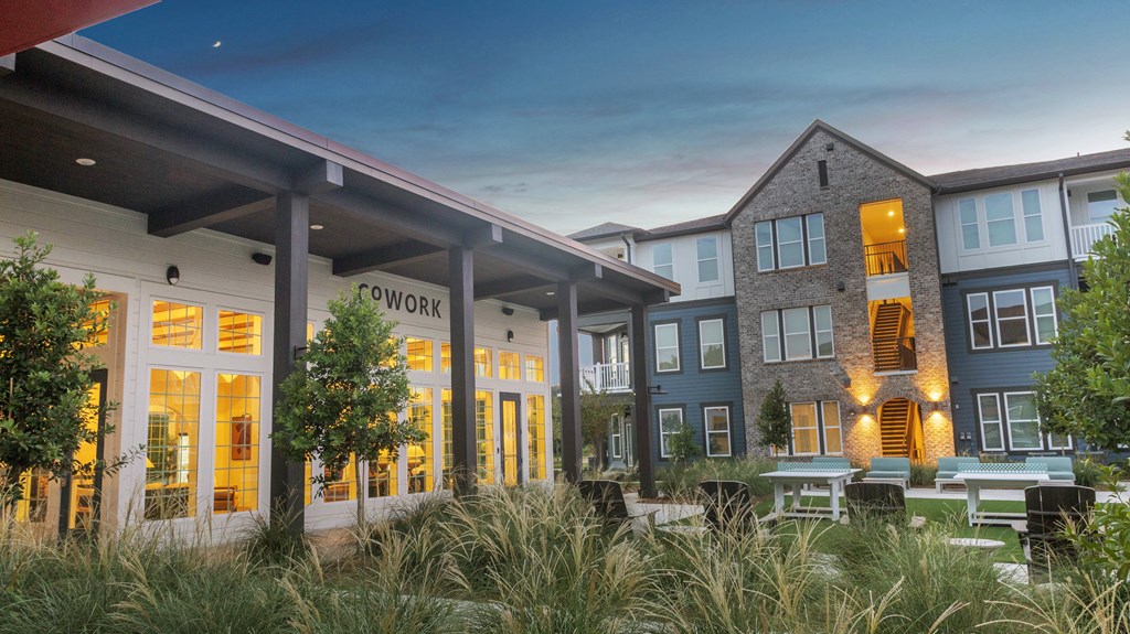 The image shows a modern building with the word "COWORK" on the facade, surrounded by greenery and set against a twilight sky.