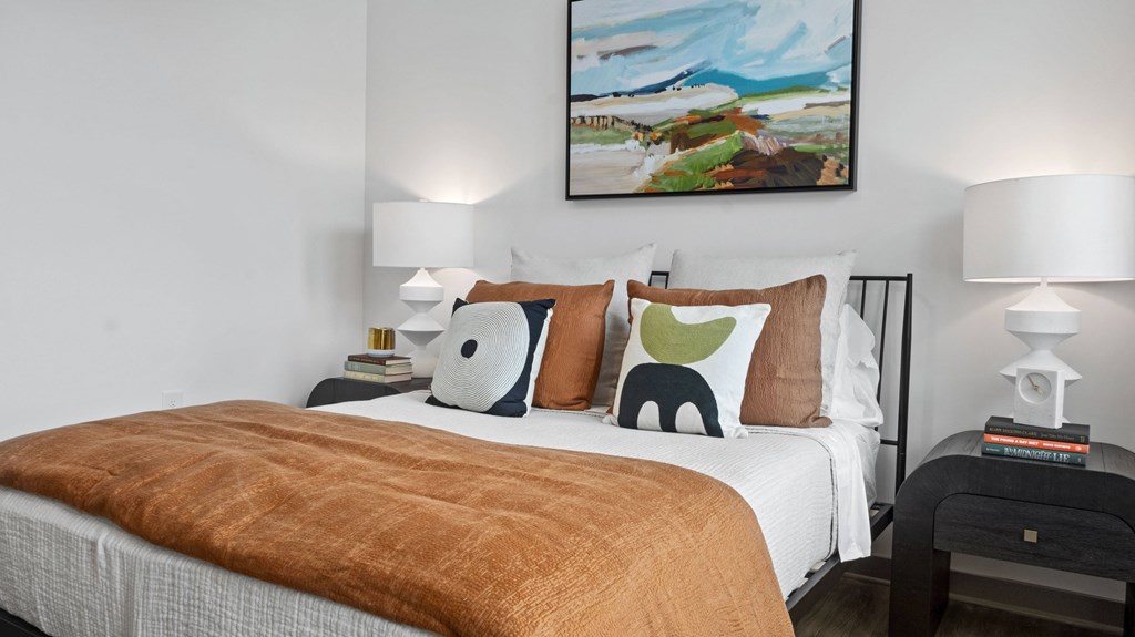 A bed with a brown blanket and white pillows is in the foreground with a painting on the wall above it.