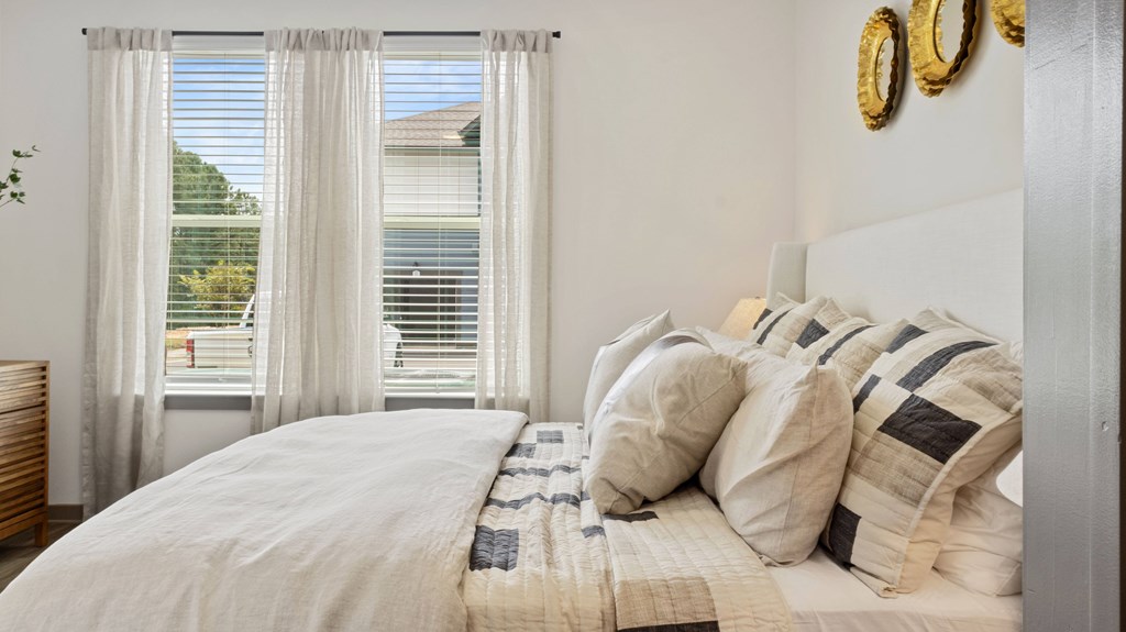 A bed with a white comforter and pillows in front of a window with white curtains.