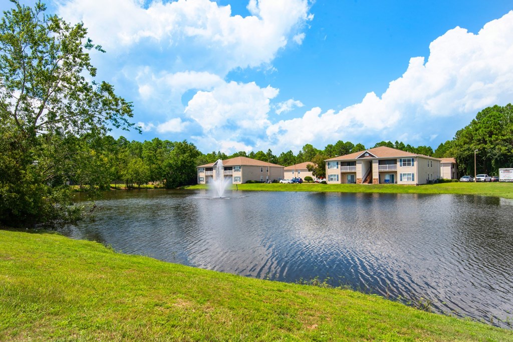 A serene pond with fountain display surrounded by greenery and apartment buildings at Crystal Lake in Pensacola, Florida.
