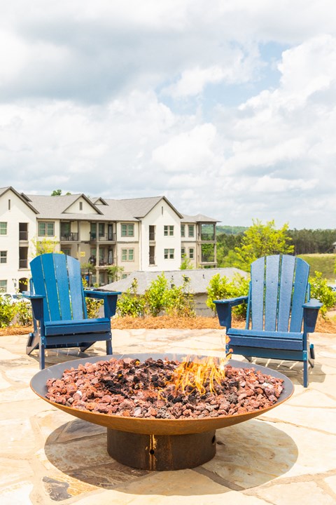 a fire pit with blue chairs on a patio with an apartment building in the background