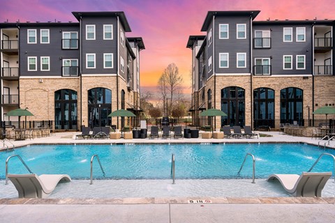 A swimming pool is surrounded by lounge chairs in front of apartment buildings.