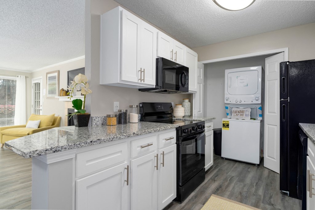 A galley-style kitchen with white cabinets, black appliances, granite countertops, and a washer and dryer in a laundry closet at Summerchase at Riverchase in Hoover, Alabama.