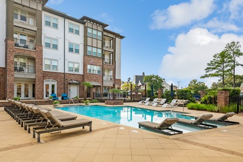 A large swimming pool surrounded by lounge chairs in front of a multi-story apartment building.