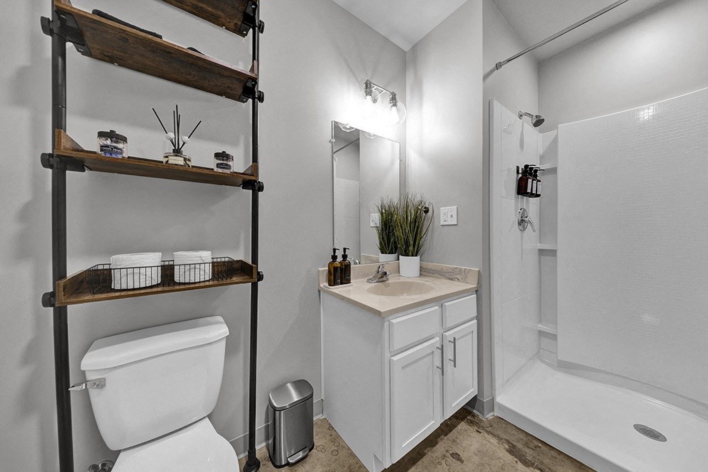Pet-Friendly Apartments In Downtown Birmingham, AL - A Bathroom With Historic Concrete Flooring, A Toilet, A Sink With White Cabinets, A Vanity Mirror, And A Shower.