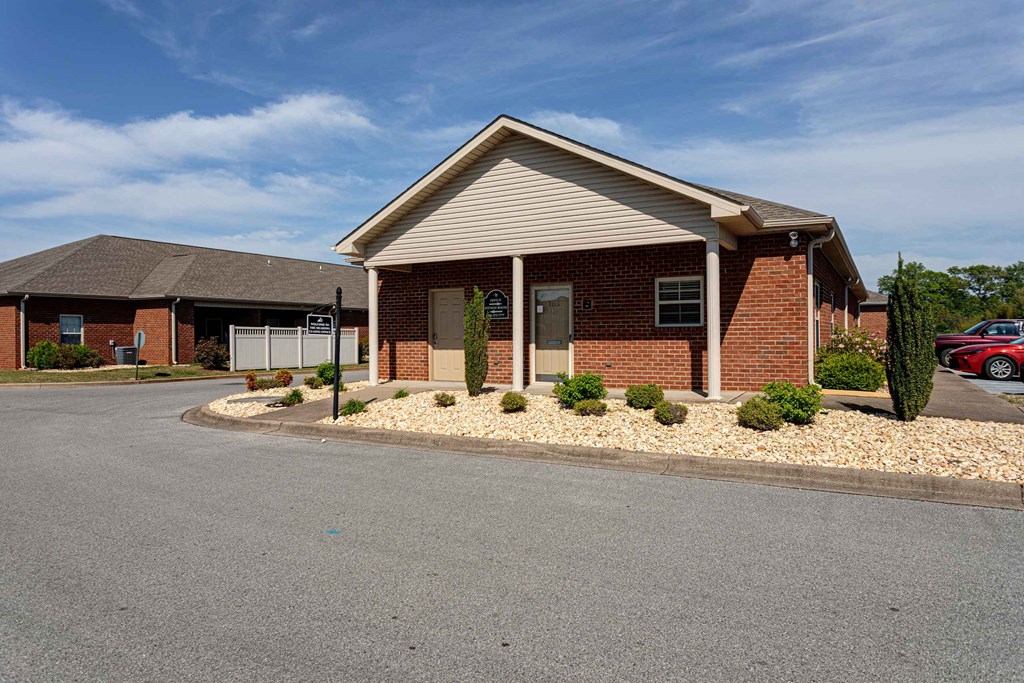 A small brick building with a gabled roof and a covered parking area in front.