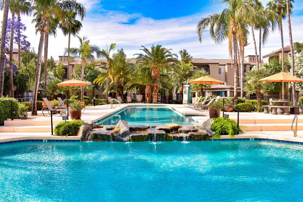 Crystal clear blue lap pool flowing over water feature into a resort-style pool surround by chaise lounge chairs, palm trees and plants