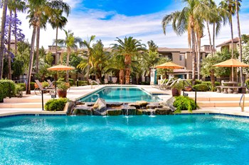 Cortina Home Page Crystal clear blue lap pool flowing over water feature into a resort-style pool surround by chaise lounge chairs, palm trees and plants - Photo Gallery 4