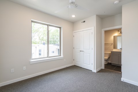 A room with a carpeted floor, a window, a door, and a bathroom visible through the doorway.