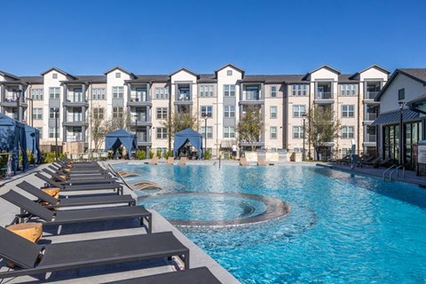A large swimming pool surrounded by sun loungers in front of apartment buildings. at Enclave on Ross Apartments, Del Valle, TX, 78617