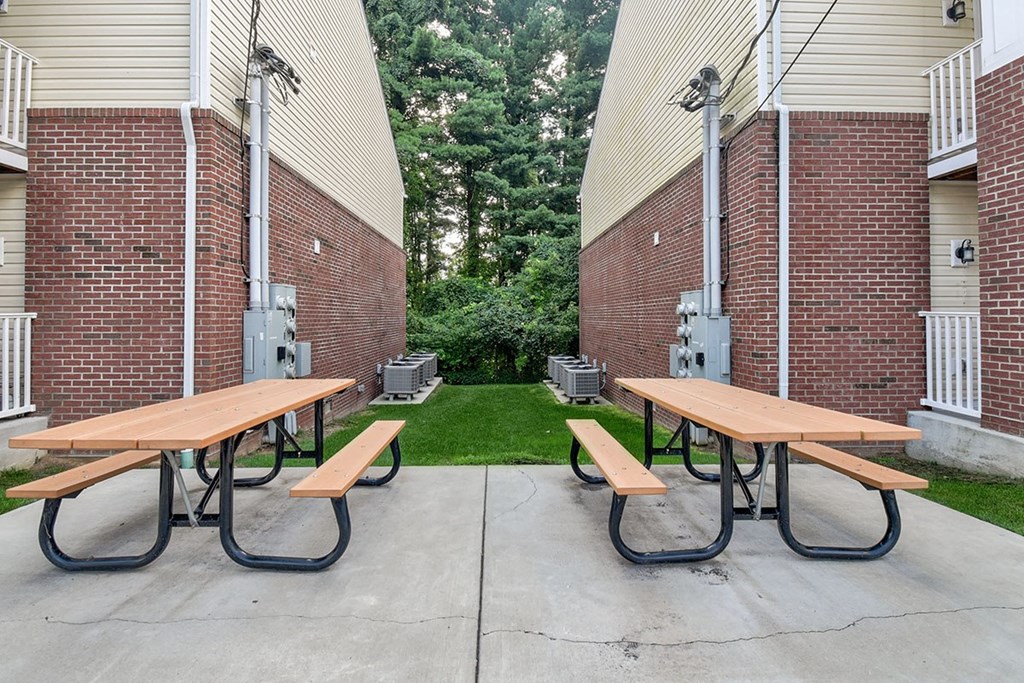 Two picnic tables are set up on a concrete patio between two brick buildings.