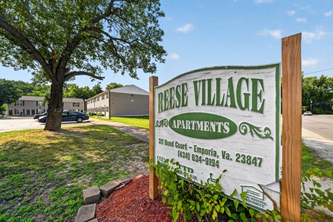A sign for Reese Village Apartments stands in front of a tree and apartment buildings.