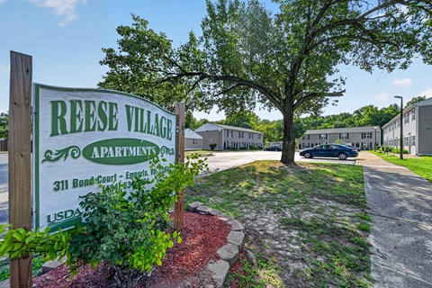 A sign for Reese Village Apartments stands in front of a tree and apartment buildings.