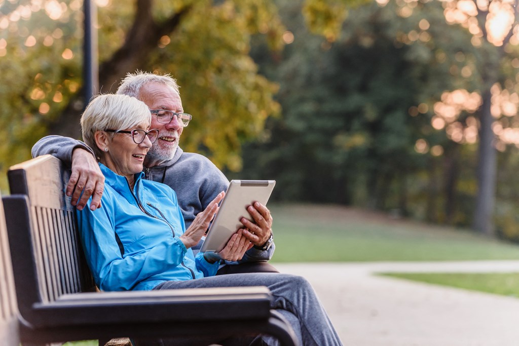 an older couple sitting on a park bench looking at a tablet