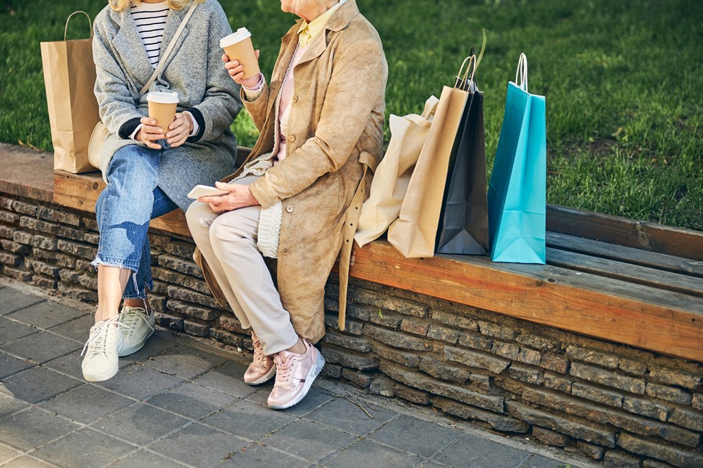 Two people sitting on a bench with shopping bags and umbrellas.