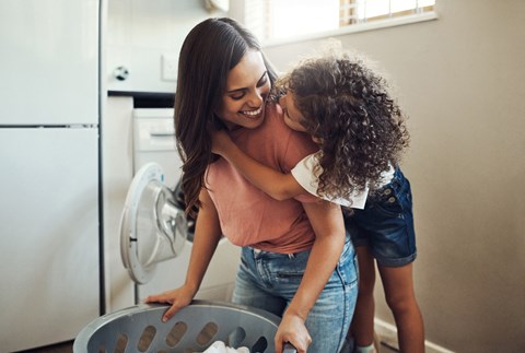 A woman and a child are standing in a kitchen, with the woman holding the child up to look into an open refrigerator.