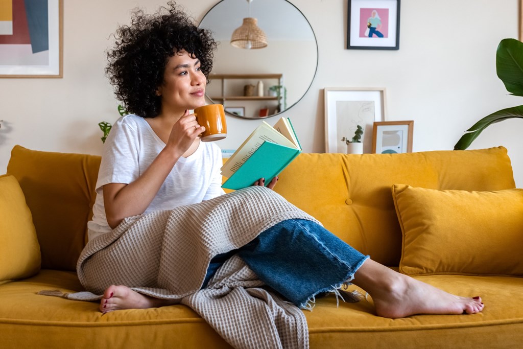 A woman with curly hair is sitting on a yellow couch, holding a mug and reading a book.