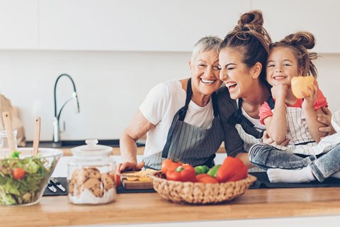 A woman and a young girl are laughing while sitting at a kitchen counter with a basket of vegetables and a bowl of salad in front of them.