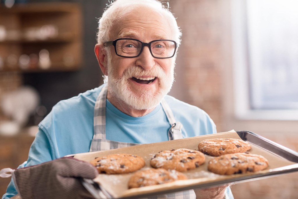 An elderly man with glasses and a beard is holding a tray of freshly baked cookies.