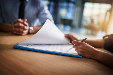 A person is writing on a notepad with a pen while another person is sitting across the table.
