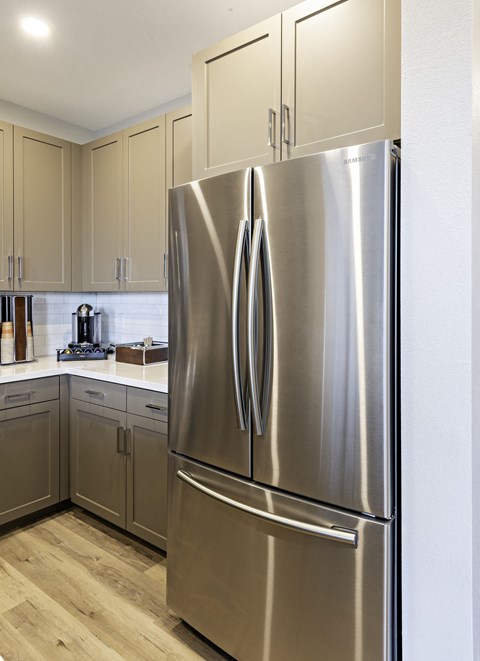 A stainless steel refrigerator stands in a kitchen with wooden floors and beige cabinets.