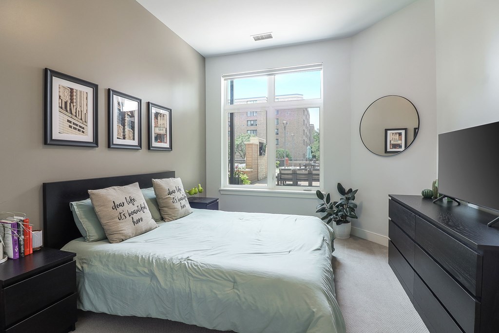 A bedroom with a bed, nightstand, and a window with a view of a building outside.