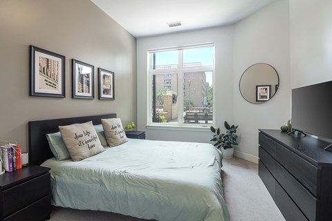 A bedroom with a bed, nightstand, and a window with a view of a building outside.