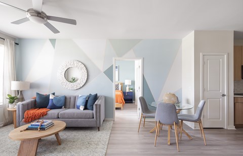 A living room with a grey sofa, a round table, a ceiling fan, and a dining table with chairs.