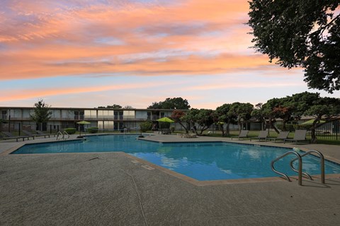 A swimming pool surrounded by trees and chairs with a sunset in the background.