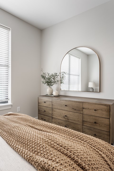 A bedroom with a bed covered in a tan blanket, a wooden dresser, and a mirror above it.