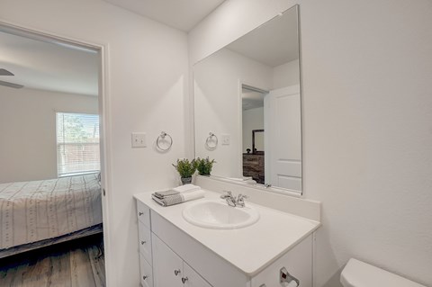 Brightly lit bathroom featuring a large mirror, white cabinets, and a seamless connection to the adjacent bedroom, offering a clean and well-integrated living space.